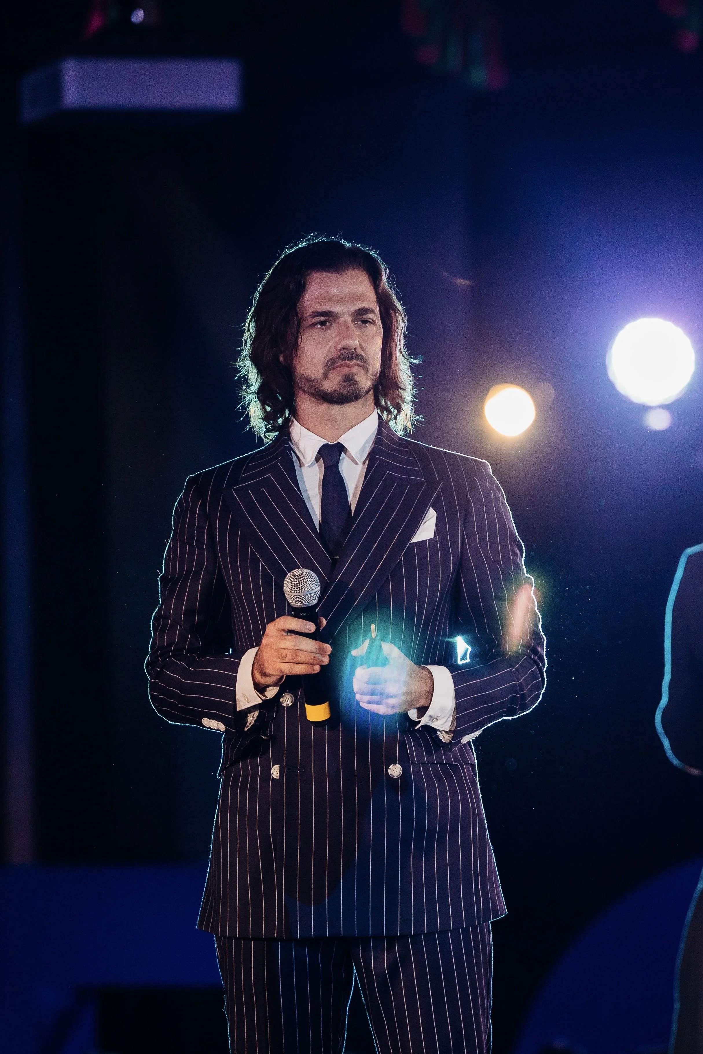 Hombre con pelo largo, barba y barba, vestido con traje a rayas, sosteniendo un micrófono en un escenario con luces traseras.