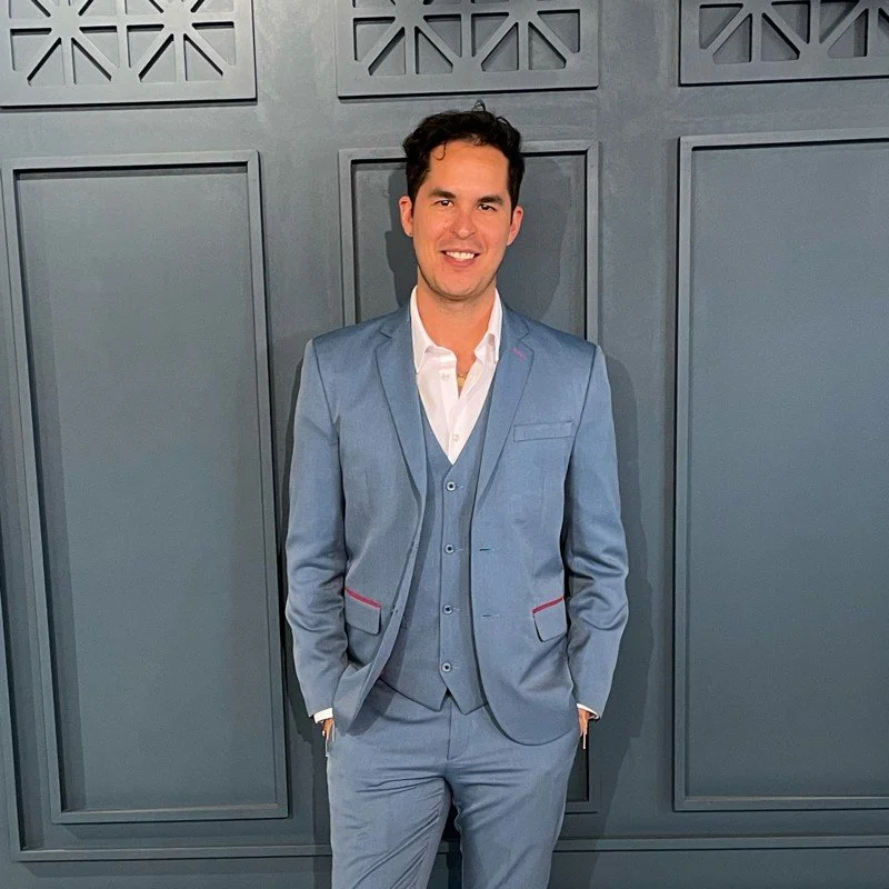 Hombre joven con traje azul claro, chaqueta y pantalones, camiseta blanca, sonriendo, de pie frente a una pared gris con detalles decorativos en las ventanas.