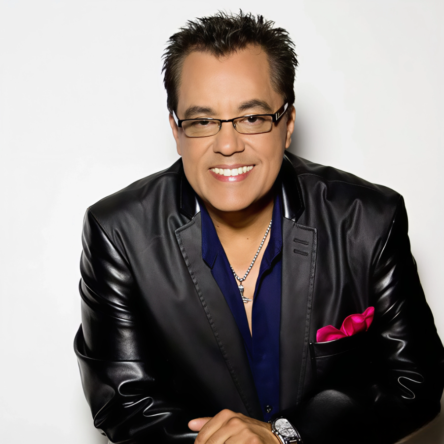 Hombre con cabello gris, vestido con saco negro, cantando con micrófono en mano, expresión emotiva.