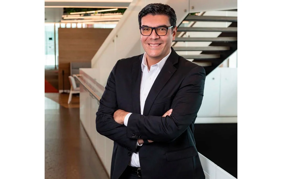 Hombre de negocios usando gafas, con traje negro y camisa blanca, sonriendo, en un ambiente moderno y luminoso.