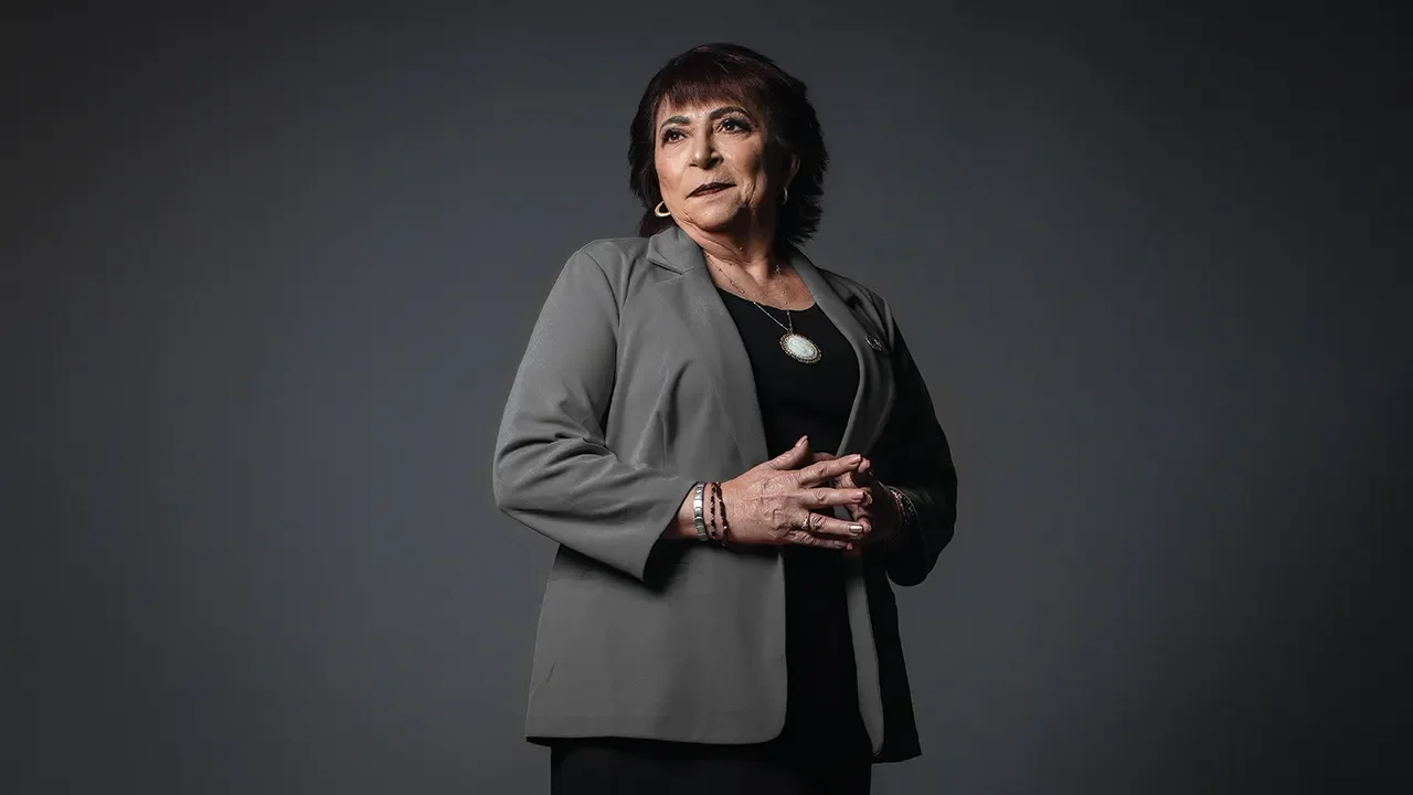 Mujer adulta de cabello corto y oscuro, vestida con blazer gris y accesorios, posando en frente de un fondo gris oscuro.