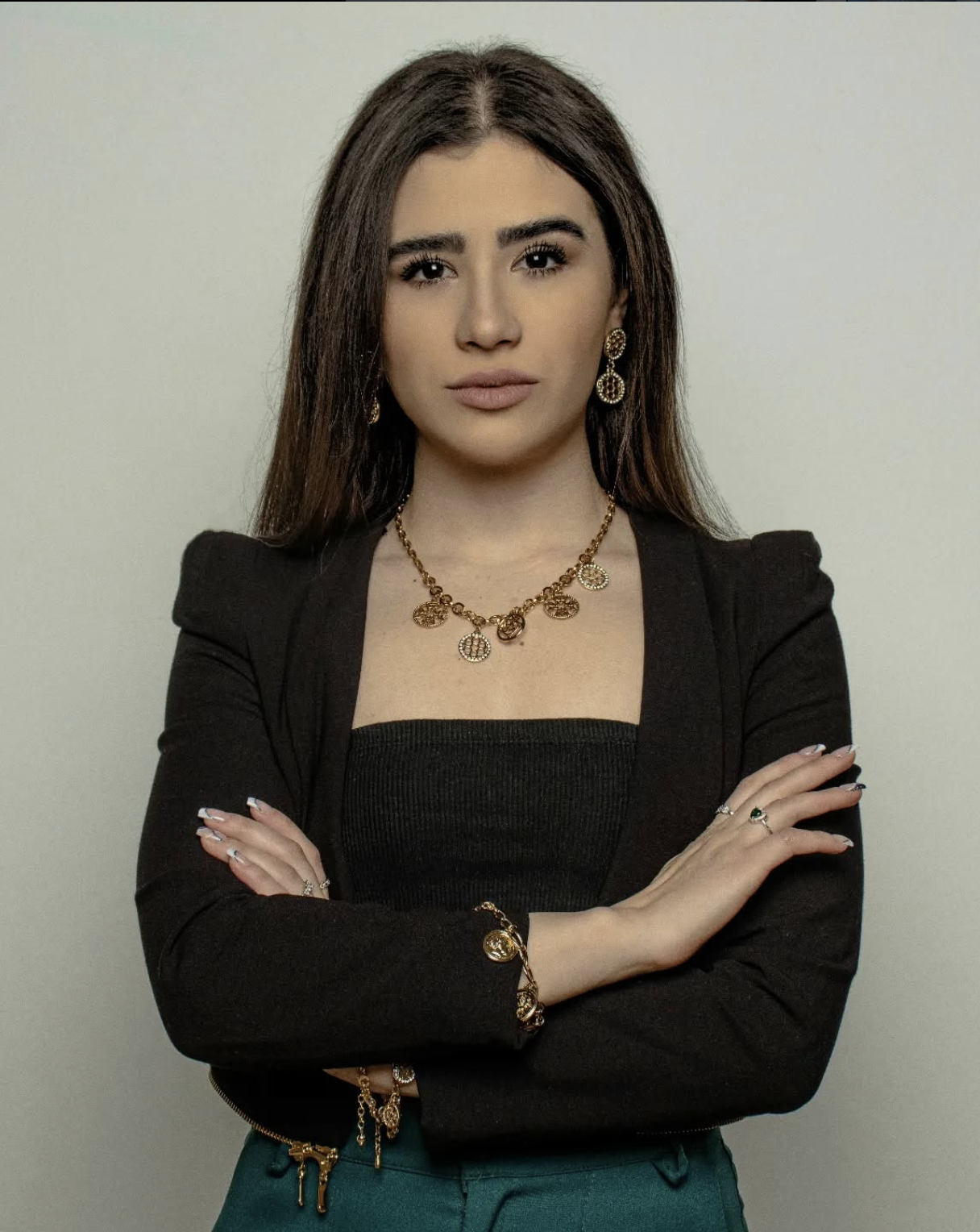 Mujer con cabello largo, vestida con blusa negra y blazer negro, usando joyería dorada, con brazos cruzados.