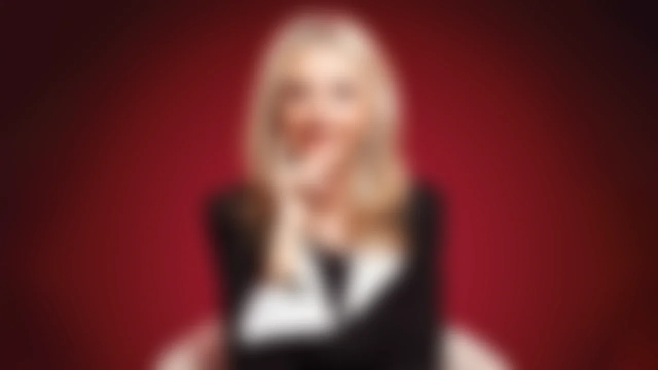 Imagen borrosa de una mujer con cabello rubio y vestido de manga larga negra con blanco, en un fondo rojo.