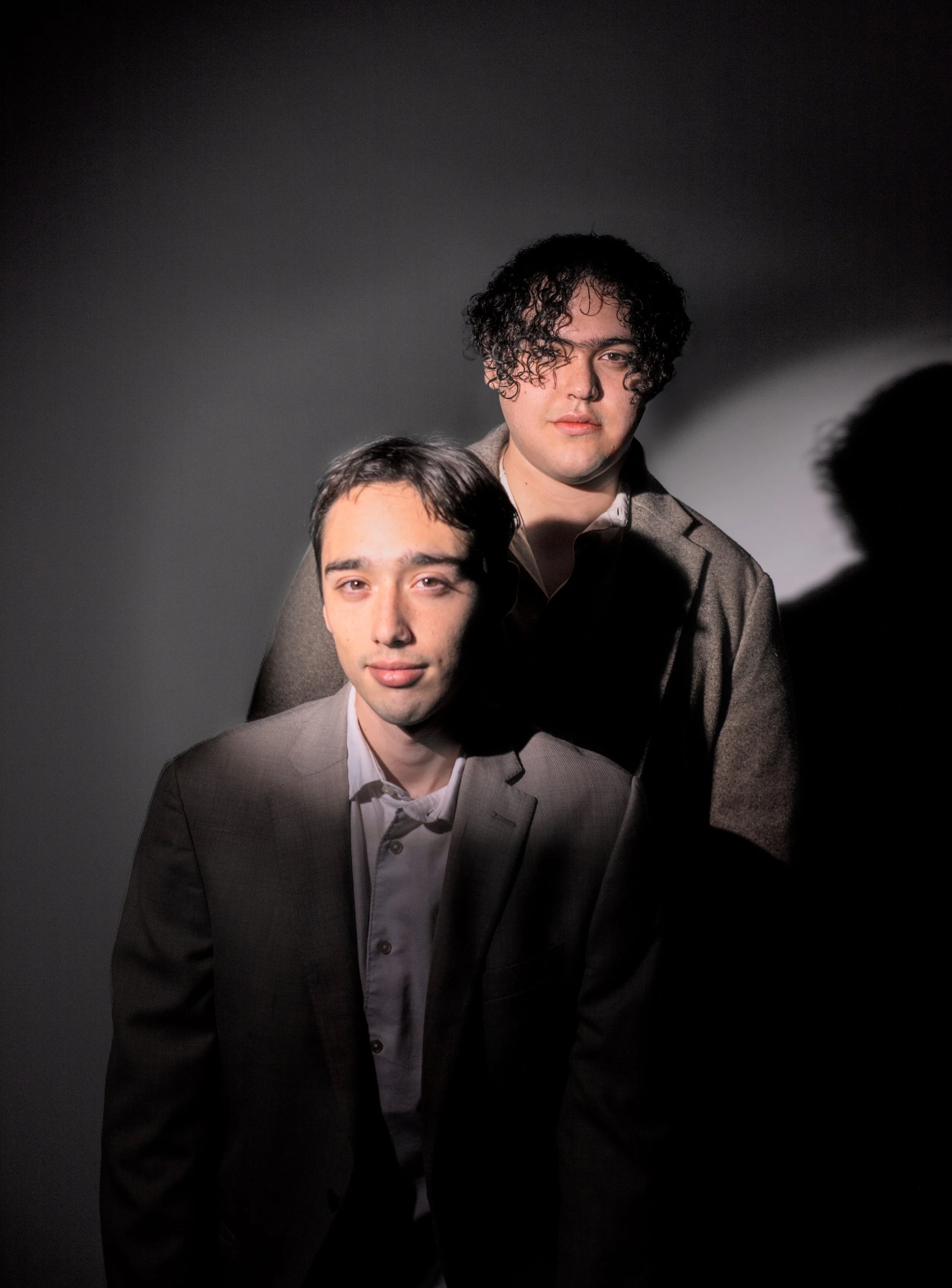 Dos hombres con trajes formales en un fondo oscuro, uno de ellos con cabello rizado y el otro con cabello liso.