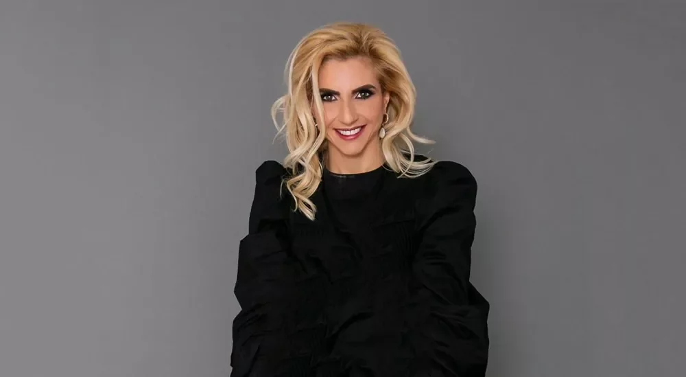 Mujer con cabello rubio y largo, sonriendo y vestida con ropa negra, de pie contra fondo gris.