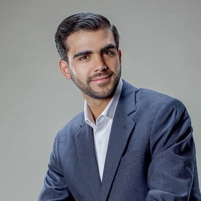 Retrato de un hombre con cabello oscuro y barba, usando un saco azul y camisa blanca, sonriendo suavemente