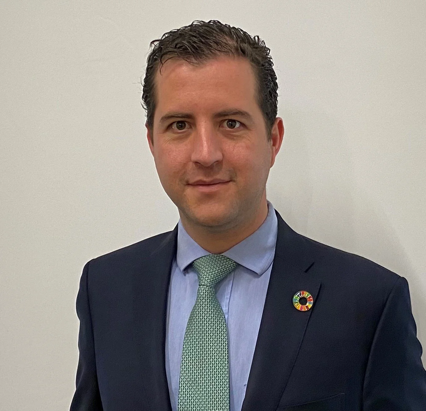Retrato de un hombre con traje oscuro, camisa azul y corbata verde, usando un pin de los objetivos de desarrollo sostenible de la ONU, frente a una pared blanca.