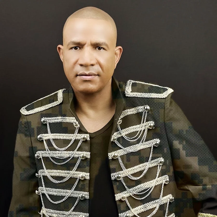 Hombre con corte de cabello calvo, usando chaqueta de militares remendada con cadenas plateadas.