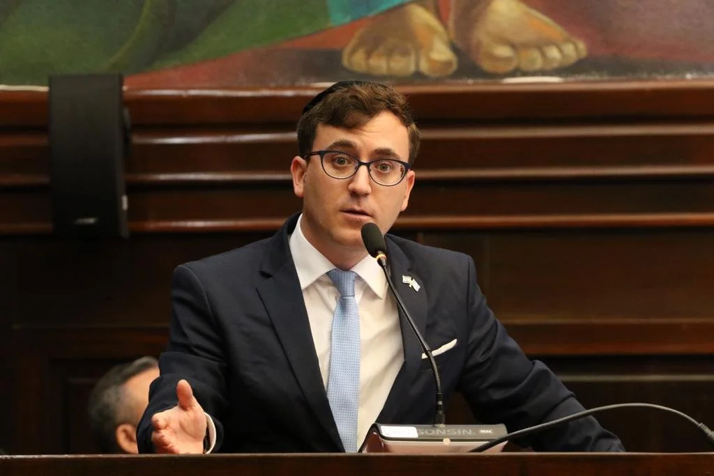 Hombre con gafas y traje oscuro hablando frente a un micrófono en una sala con fondo de madera y pintura en la pared.