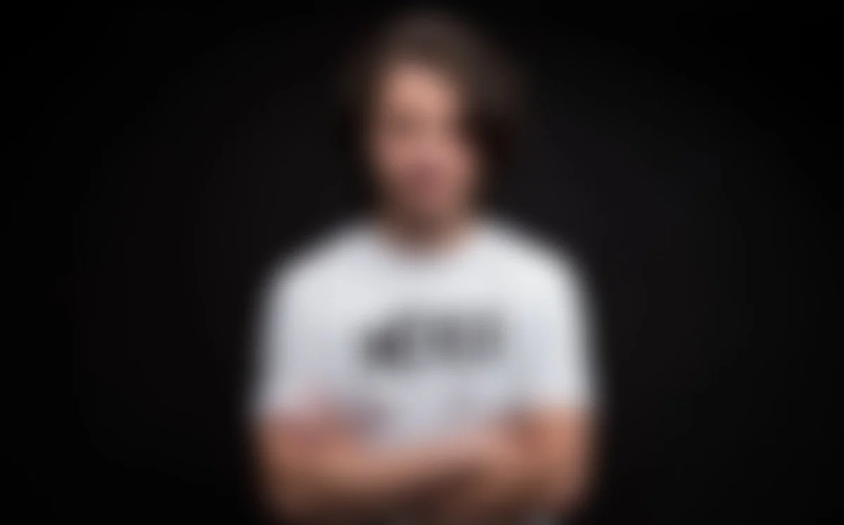 Persona con camiseta blanca frente a fondo negro, la imagen está desenfocada.