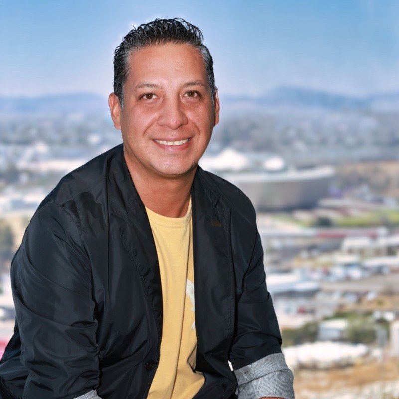 Hombre sonriente con chaqueta negra y camiseta amarilla, fondo de ciudad y montañas.