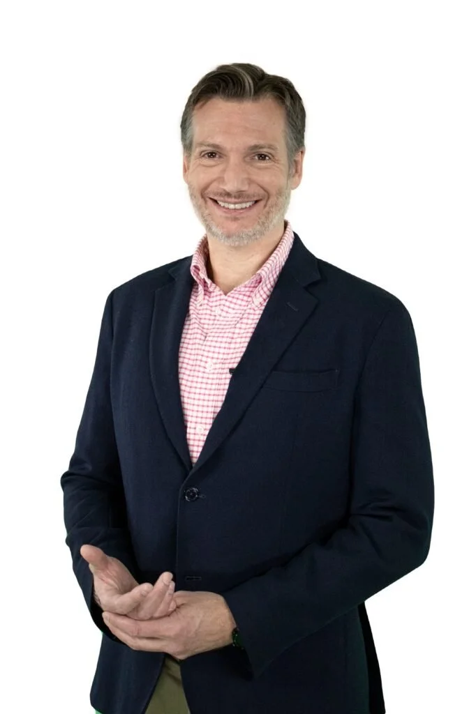 Hombre sonriendo, usando saco azul y camisa a cuadros rosa en fondo blanco.