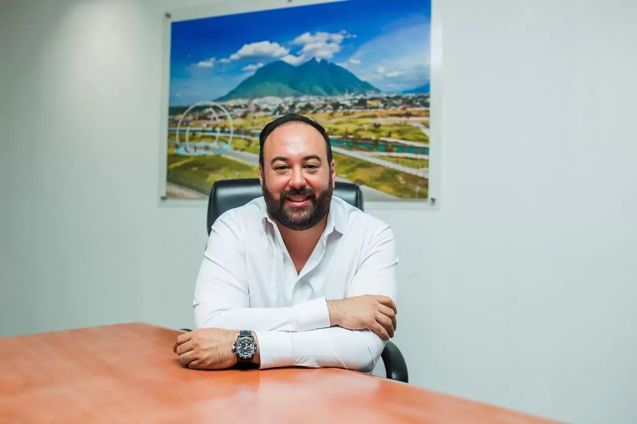 Hombre sonriendo sentado en una oficina con una pintura de un paisaje montañoso de fondo.