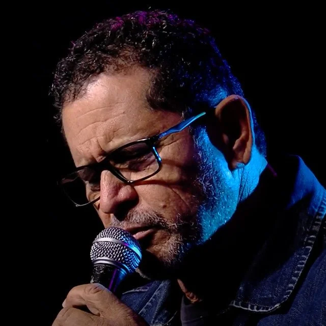 Hombre con gafas y barba, cantando o hablando en un micrófono, iluminado con luces azules y rosas en un fondo oscuro.