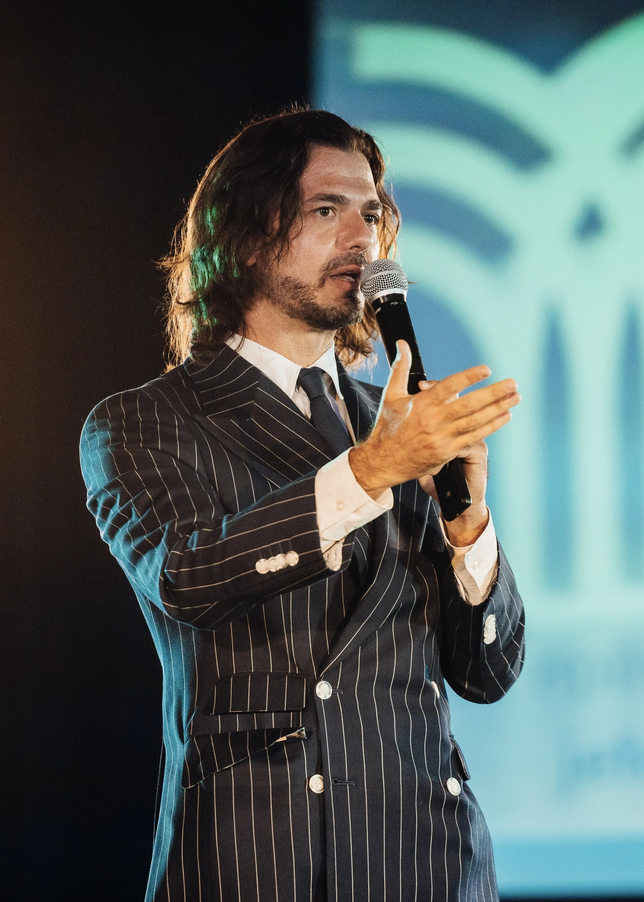 Hombre con cabello largo, barba y vestido a rayas, hablando con micrófono en mano en un escenario.