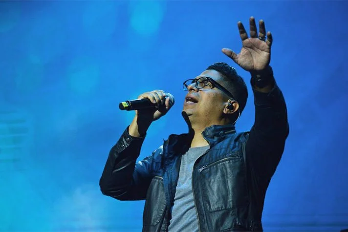 Hombre cantando en un escenario con micrófono, usando gafas, chaqueta negra y fondo azul.