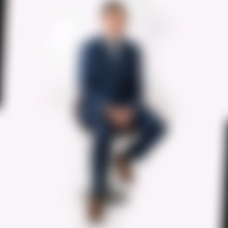 Persona sentada en una silla blanca contra un fondo blanco, llevando ropa oscura, con cabello rubio y peinado en pixie.