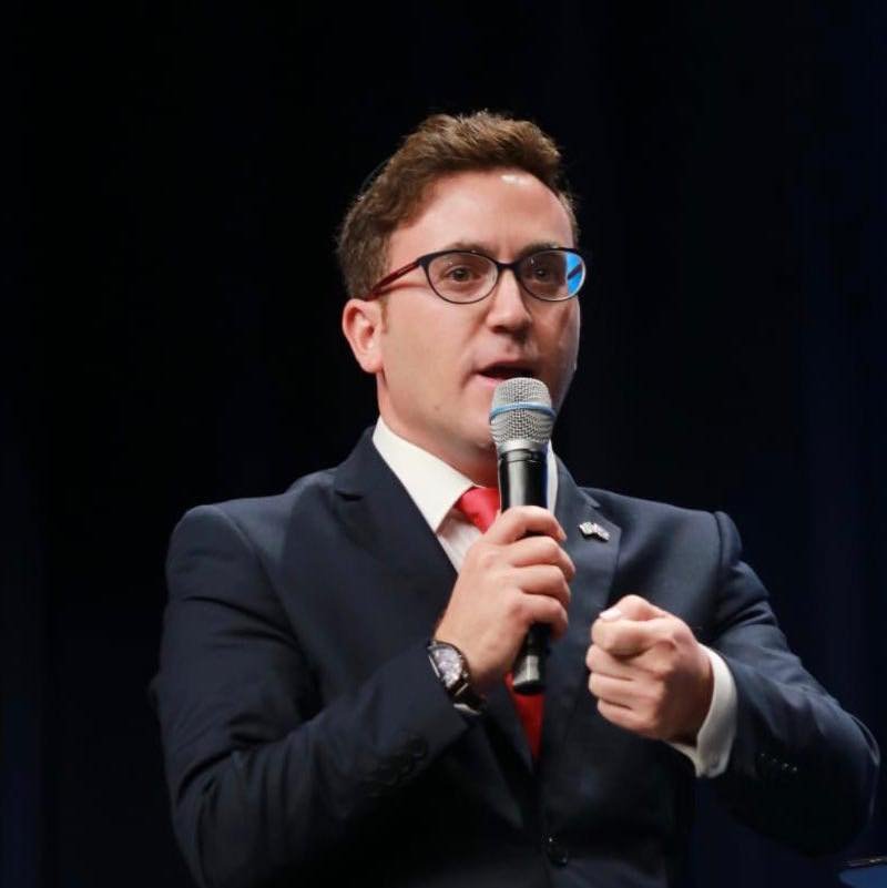 Hombre con gafas, traje y corbata hablando en un micrófono en una sala de reuniones. Panel de madera y pintura de pies en el fondo.