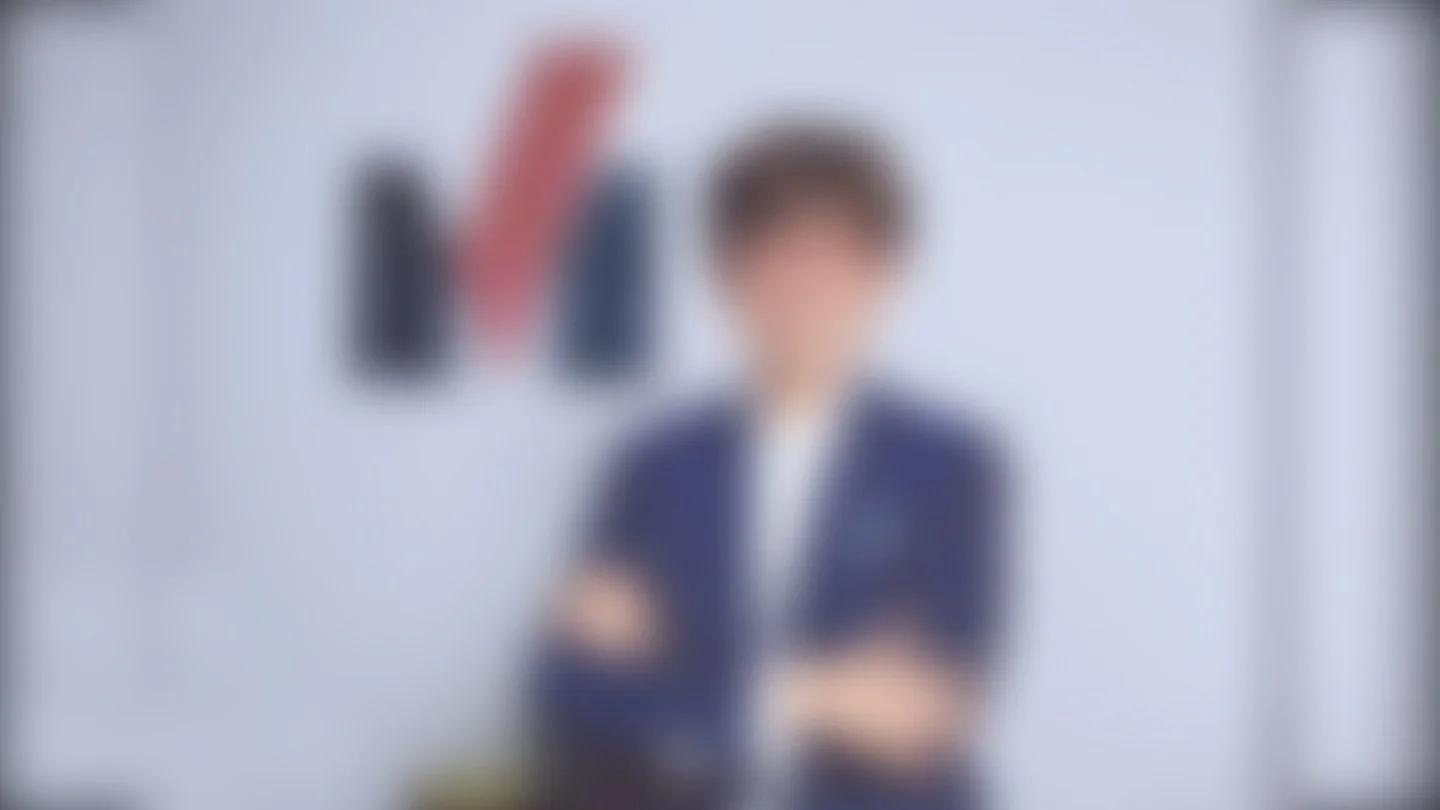 Persona de pie con fondo de libros en una pared blanca, vestida con camisa blanca y chaqueta azul oscuro.