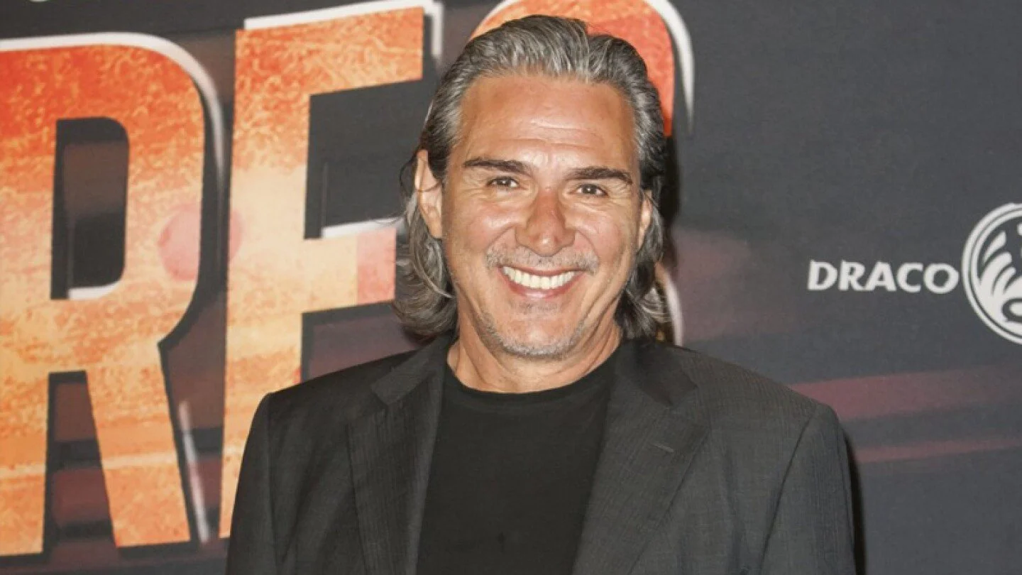 Un hombre con cabello gris recogido y barba, vestido con una chaqueta negra y camiseta negra, sonriendo frente a un fondo con letras y logotipos.
