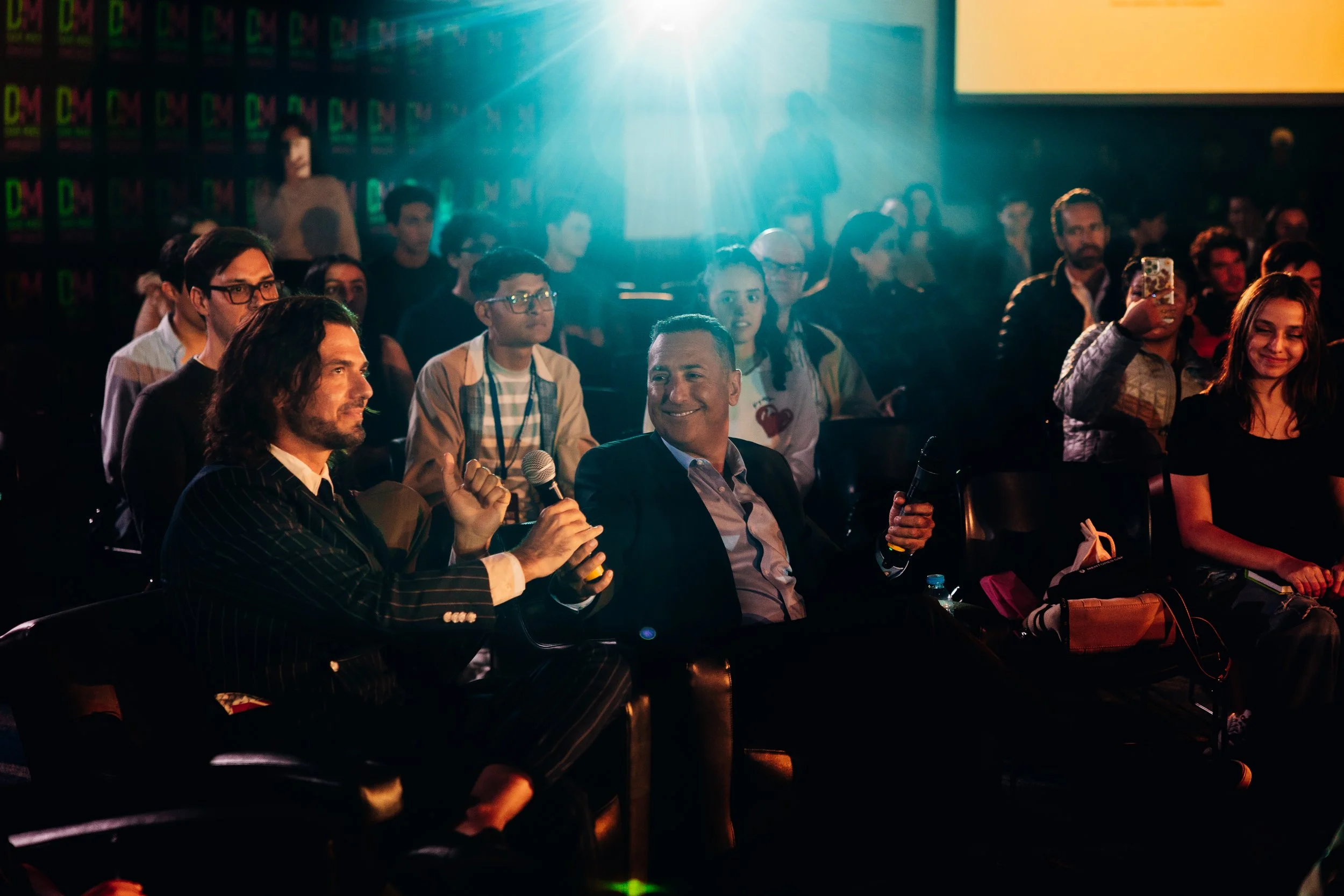 Personas en un evento, con luces brillantes, algunas tomando fotos y otras conversando, en un ambiente de conferencias o presentaciones.