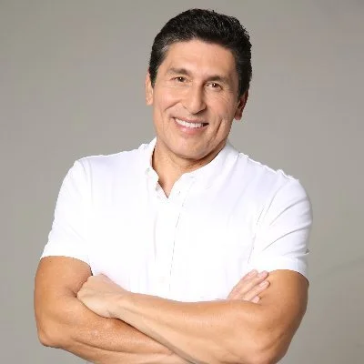Hombre con cabello negro, sonriente, usando una camiseta blanca, con los brazos cruzados, fondo gris.