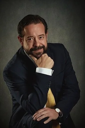 Hombre con barba, vestido con traje oscuro y corbata amarilla, posando con la mano en la barbilla