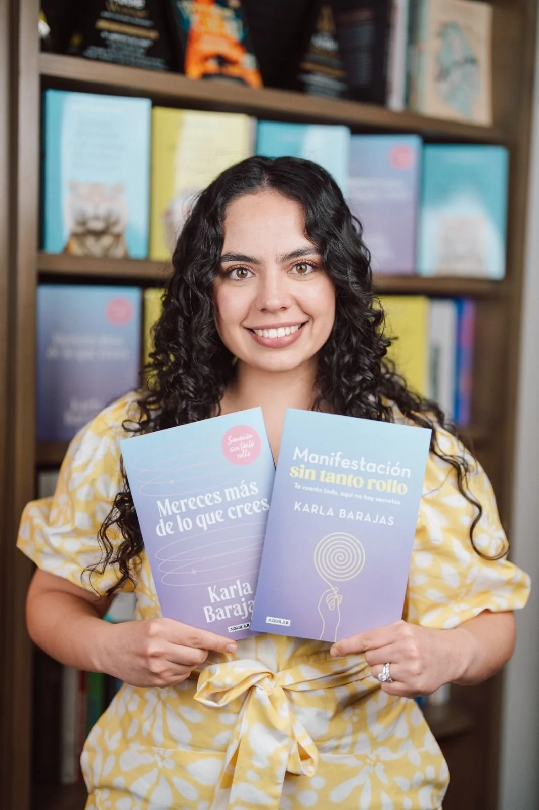 Una mujer con cabello rizado y vestido amarillo sosteniendo dos libros, uno titulado 'Mereces más de lo que crees' y otro 'Manifestación sin tanto rollo', en una librería con estantes llenos de libros.
