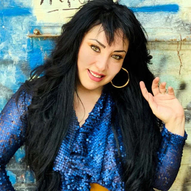 Mujer con cabello largo y oscuro, haciendo un gesto con la mano, usando un vestido azul brillante y un aro grande, frente a una pared pintada de azul y beige.