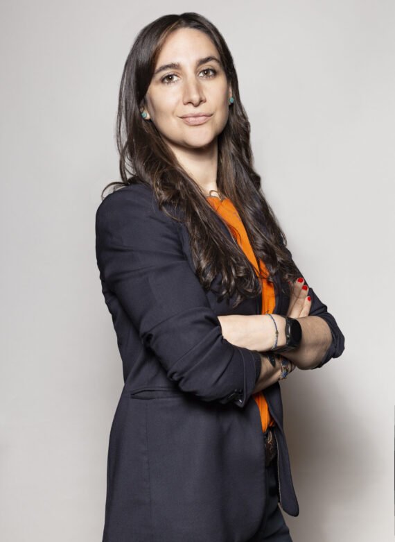 Retrato de una mujer con cabello largo y oscuro, usando una chaqueta negra y una blusa naranja, cruzada de brazos, posando contra un fondo gris claro.
