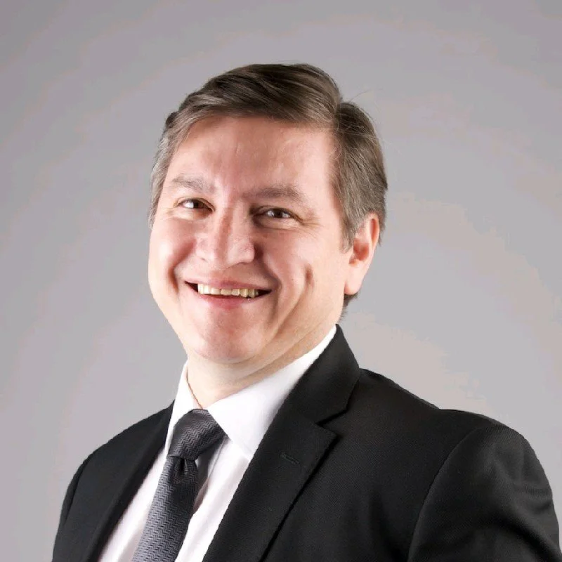 Hombre sonriendo, vestido con traje negro y corbata, fondo gris claro.