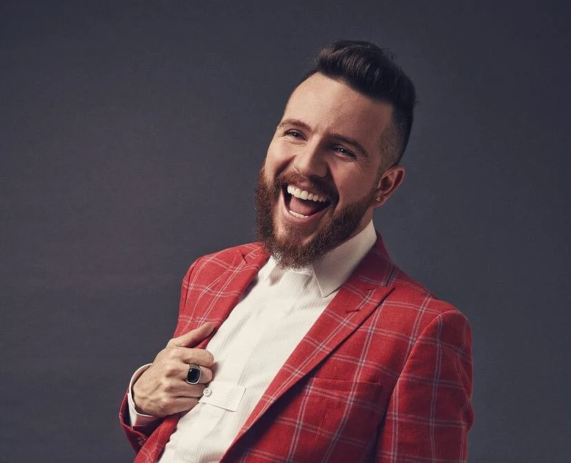 Hombre de barba y peinado moderno, sonriendo ampliamente, vestido con una americana roja de cuadros y una camisa blanca, en un fondo gris oscuro.