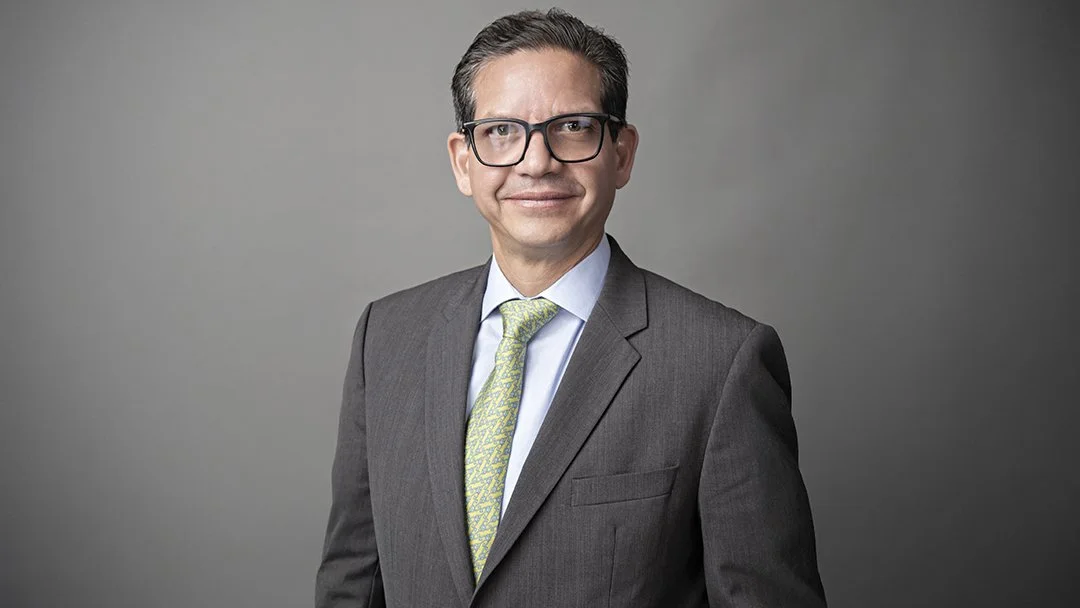 Hombre con gafas y traje gris, camisa celeste y corbata de color verde, sonriendo en un fondo gris.
