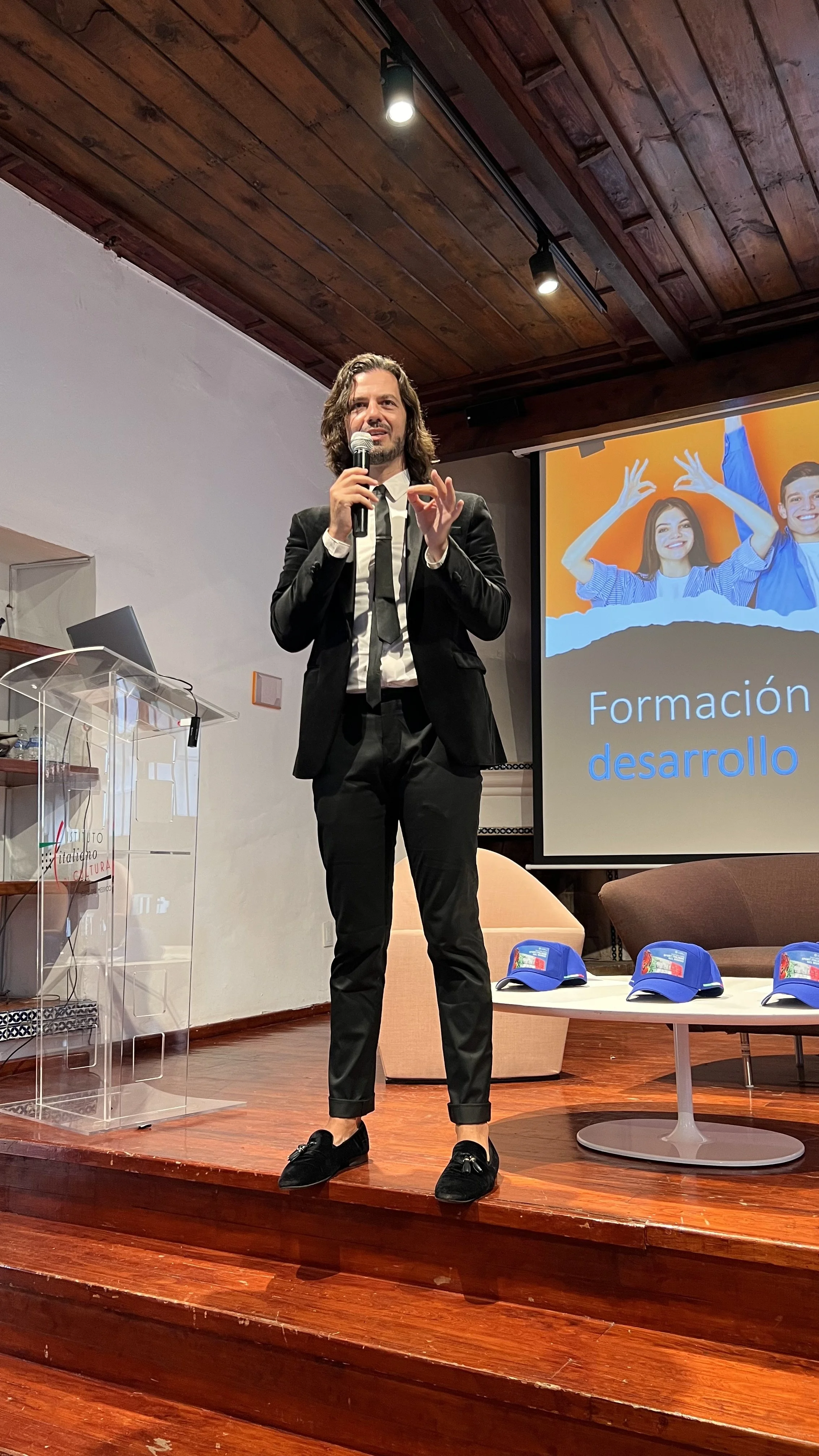 Hombre con barba y cabello largo hablando en una conferencia, vestimenta formal en un escenario de madera, con una pantalla que dice 'Formación desarrollo', en un entorno interior con sillones y gorras en la mesa.