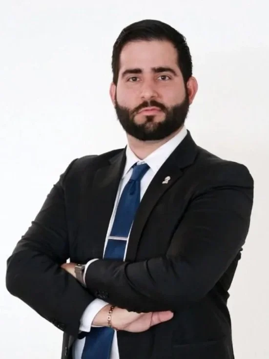 Hombre con barba, vestido con saco negro, camisa blanca, corbata azul y con los brazos cruzados frente a una pared blanca.