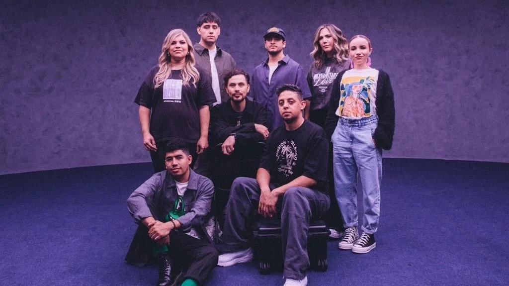 Grupo de ocho jóvenes posando en un fondo gris oscuro con iluminación suave.