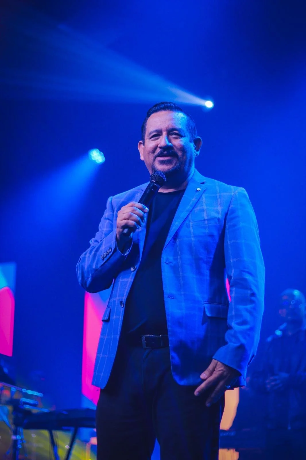 Hombre con cabello oscuro y bigote, vestido con traje de rayas y corbata plateada, en un fondo azul.