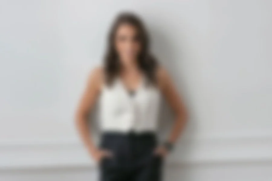 Mujer de pie con camiseta blanca y pantalones oscuros, posando con las manos en los bolsillos frente a una pared gris.