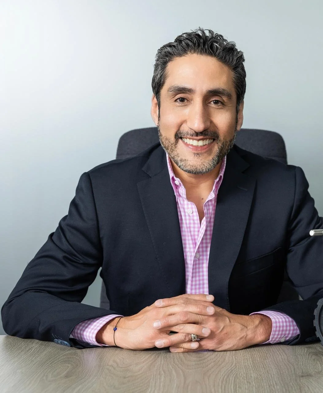 Hombre de complexión media con barba, cabello oscuro y gris, sonriendo, sentado en una silla de oficina, vestido con saco negro y camisa rosa con cuadros blancos, en un ambiente de oficina con fondo neutro.