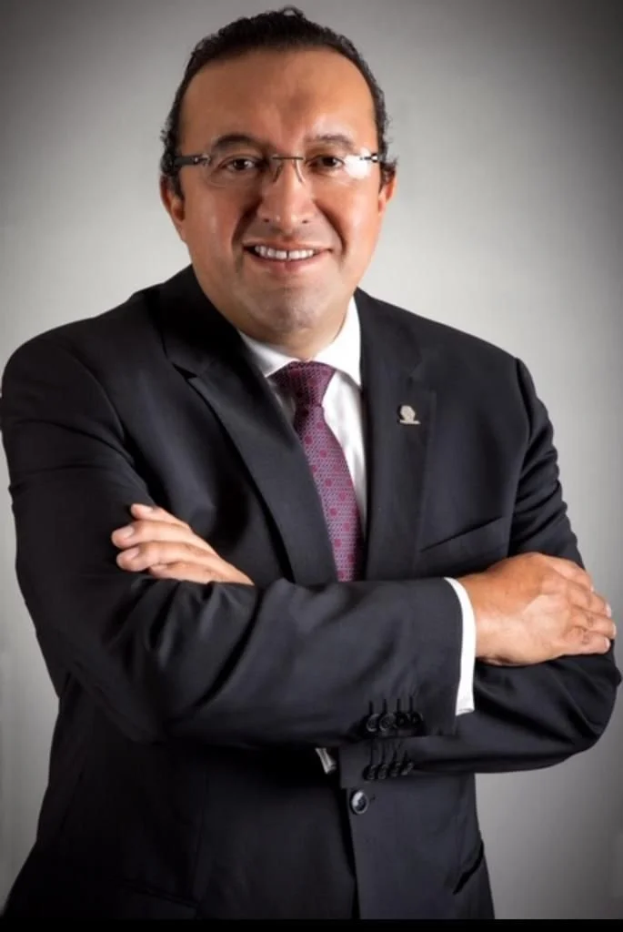 Retrato de un hombre de negocios con traje, corbata y gafas, sonriendo y con los brazos cruzados frente a un fondo gris.
