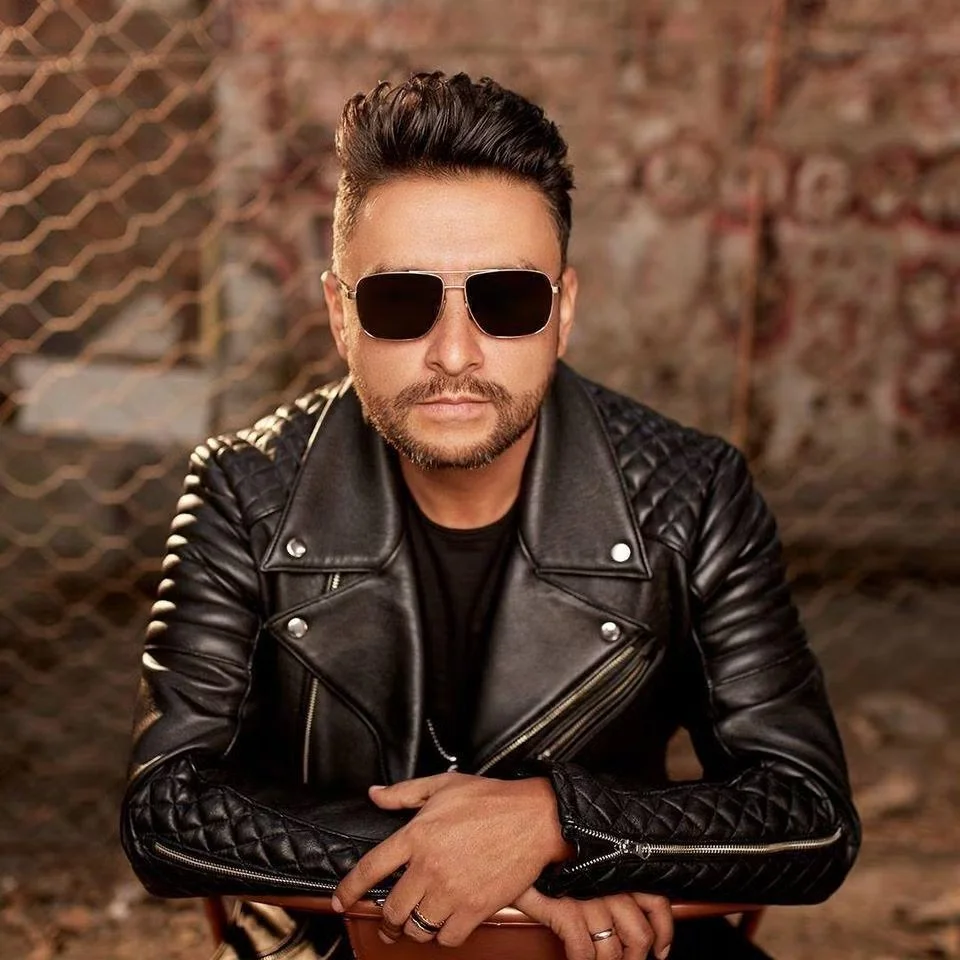 Hombre con cabello oscuro y barba, usando gafas de sol oscuras, chaqueta de cuero negro con detalles de costuras acolchadas en las mangas, en un entorno con fondo de pared de ladrillos y malla metálica.