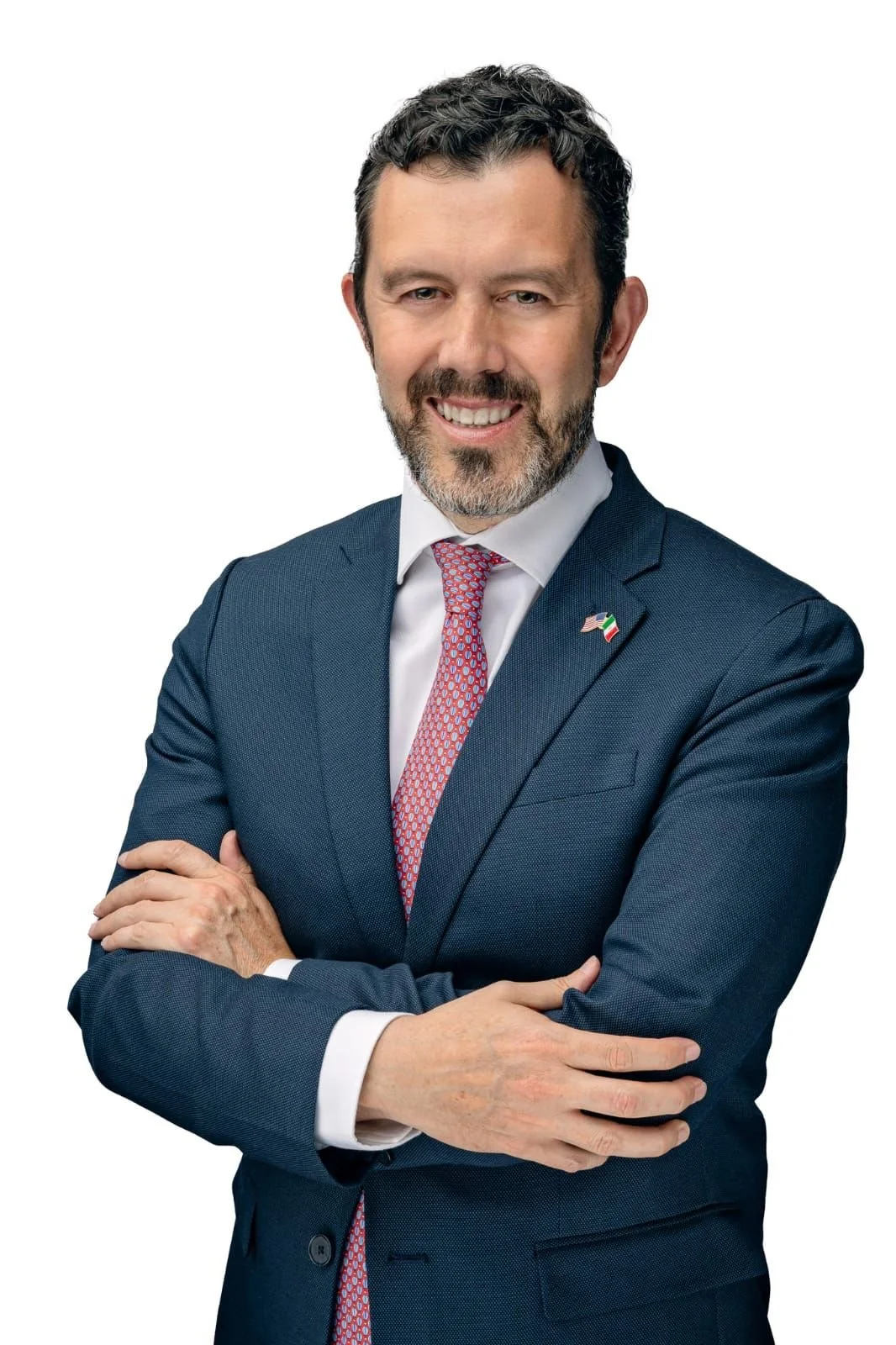 Hombre de negocios con barba, vestir de traje azul, camisa blanca, y corbata rosa, sonriendo con los brazos cruzados en fondo blanco.