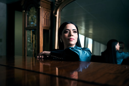 Mujer de cabello oscuro, vestida con ropa azul, descansando su brazo en una mesa de madera, en un entorno interior con ventanas y otra persona al fondo.