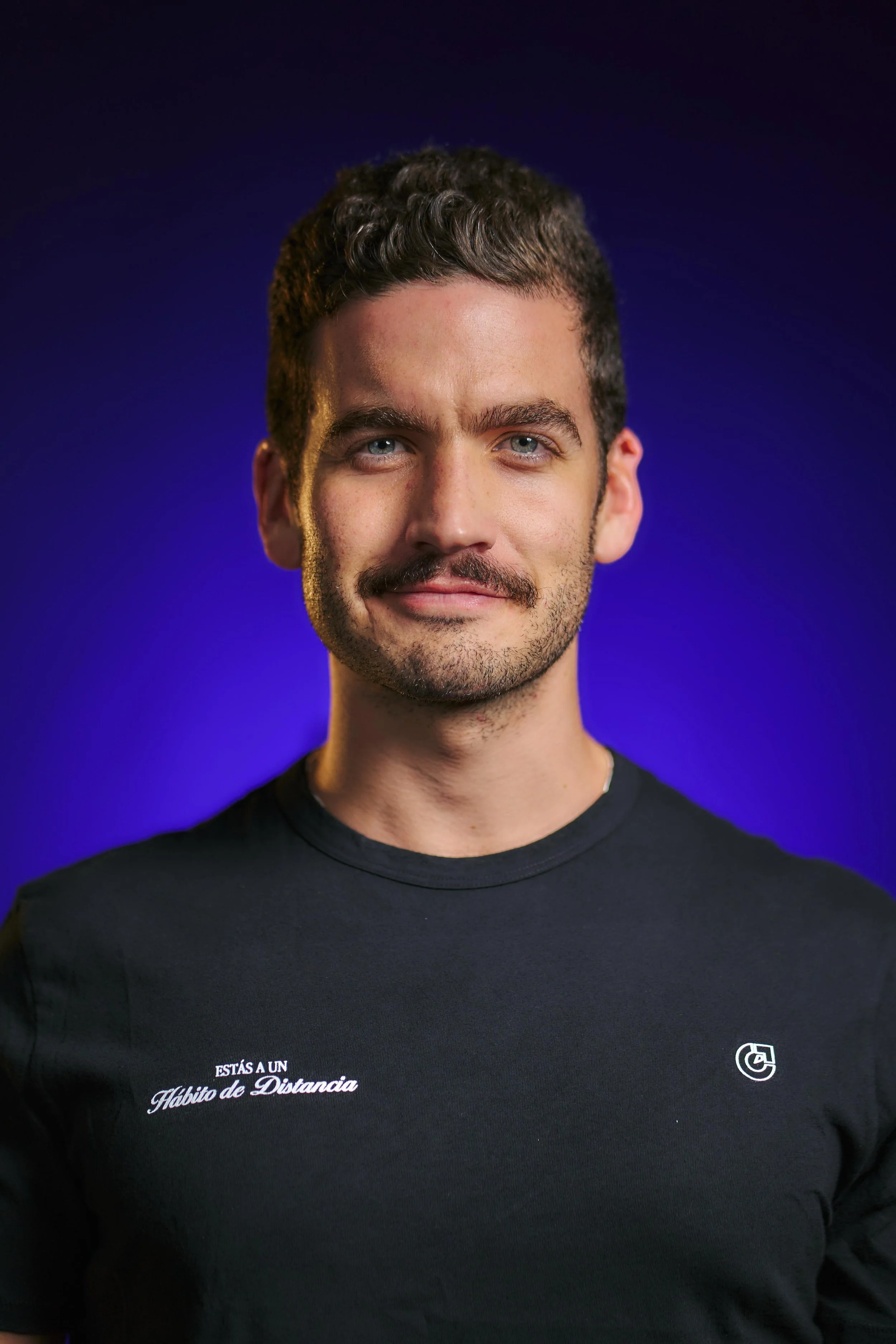 Persona con cabello castaño en un fondo oscuro, sonriendo y vestida con camiseta blanca.