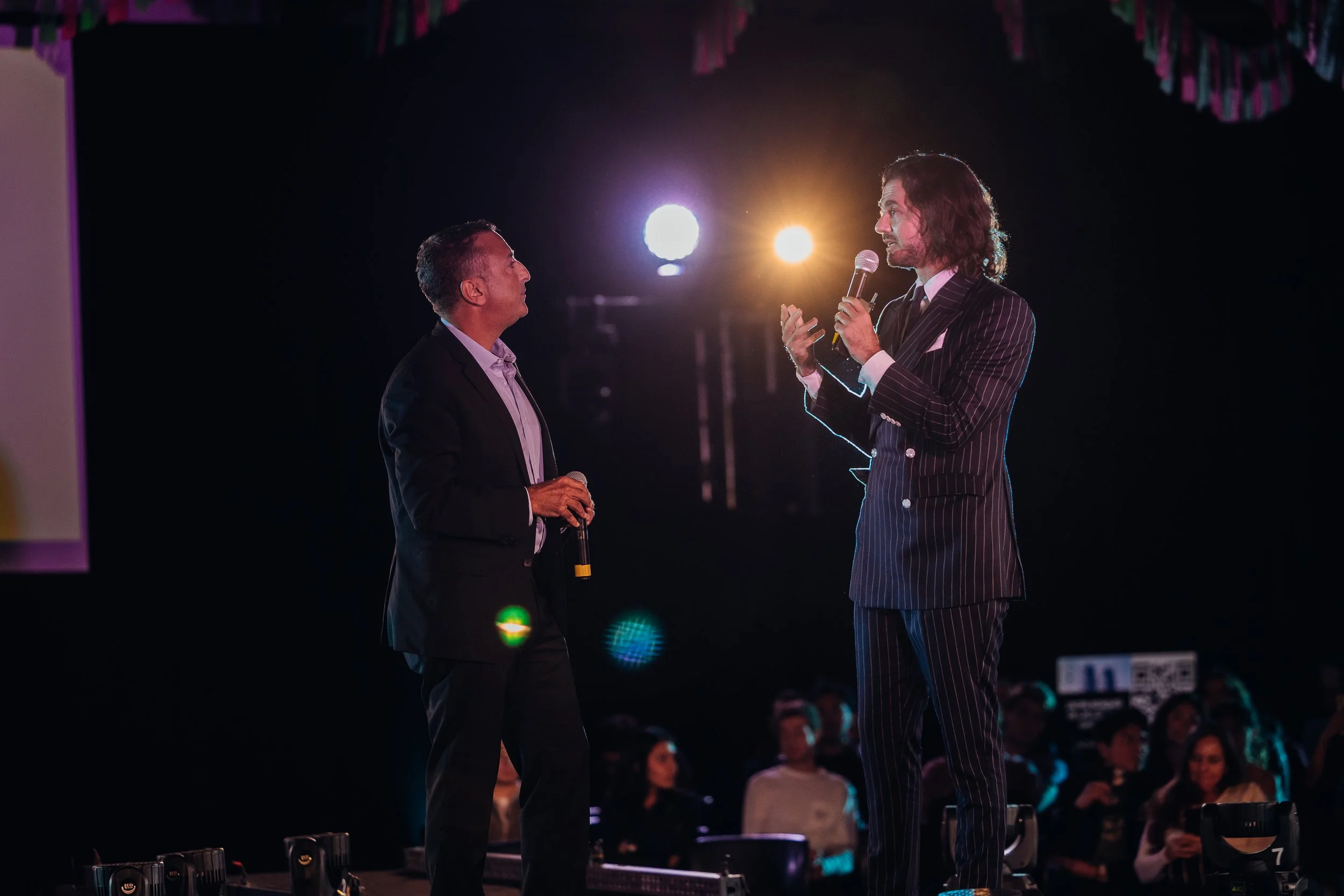 dos hombres de pie en un escenario con micrófonos, hablando frente a un público, con luces brillantes detrás.