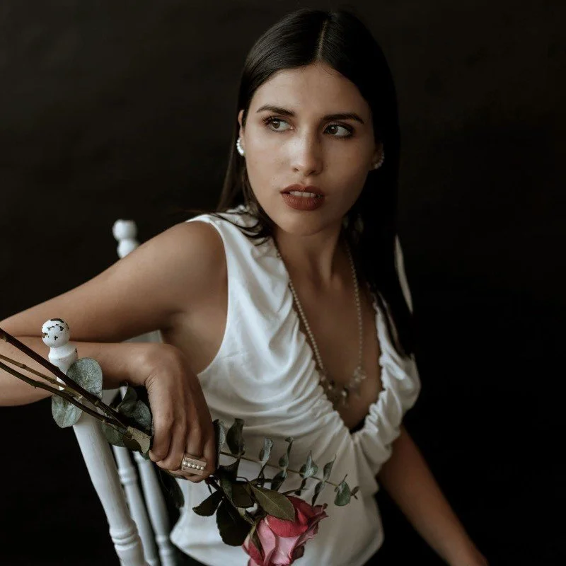 Mujer vestida con blusa blanca y accesorios, sosteniendo una rosa y mirando hacia un lado.
