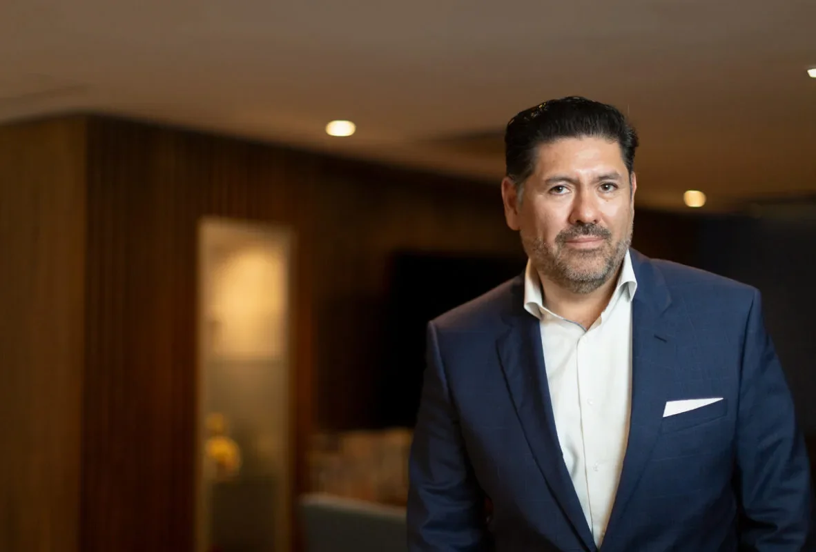 Hombre con barba, cabello oscuro y peinado hacia atrás, vestido con traje azul y camisa blanca, posando en un ambiente interior.