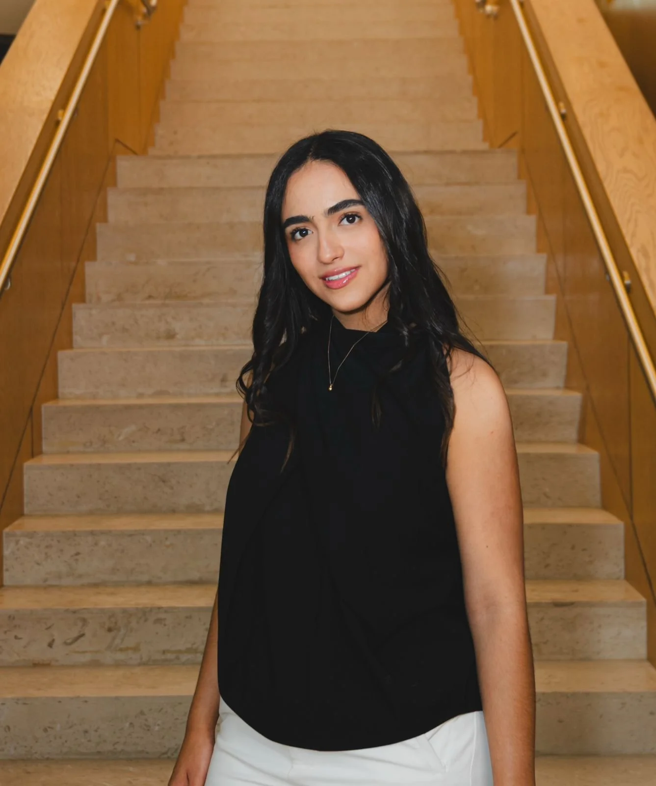 Mujer joven con cabello oscuro y rostro sonriente, vestida con blusa negra y pantalones blancos, de pie en una escalera de madera.