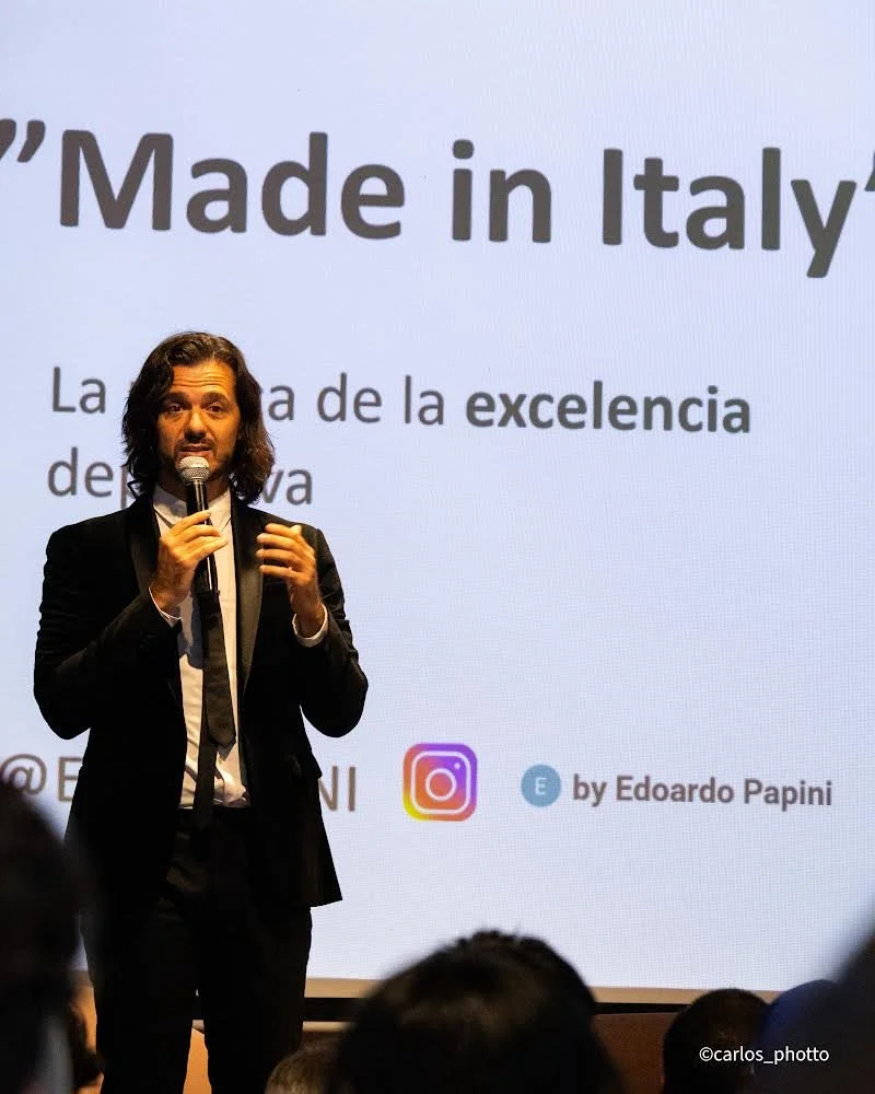 Hombre de cabello largo y barba, vestido con traje negro, hablando en un micrófono frente a una pantalla que dice "Made in Italy" y otras palabras en español, en un evento o conferencia.