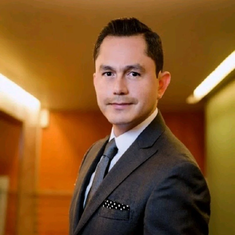 Hombre con cabello oscuro peinado hacia atrás, vestido con traje gris, camisa blanca y corbata gris, en un ambiente con paredes de madera y luz cálida.